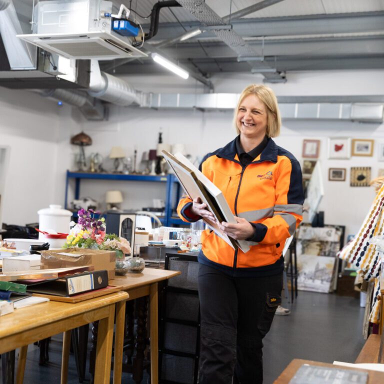 A woman working in one of Bristol Waste's Reuse Shops. She is wearing an orange zip-up jacket with the Bristol Waste logo on it. She is holding some large photo frames. She is walking and smiling. In the background is lots of other items, shelving units, and a big wooden table.