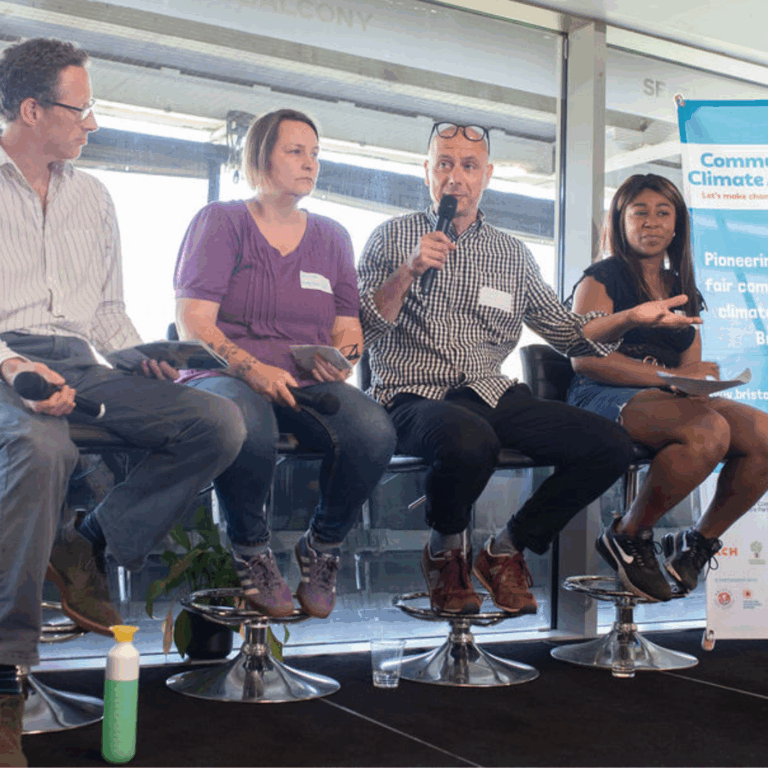 Four people sitting on a stage. One person is holding a microphone and speaking to the audience.