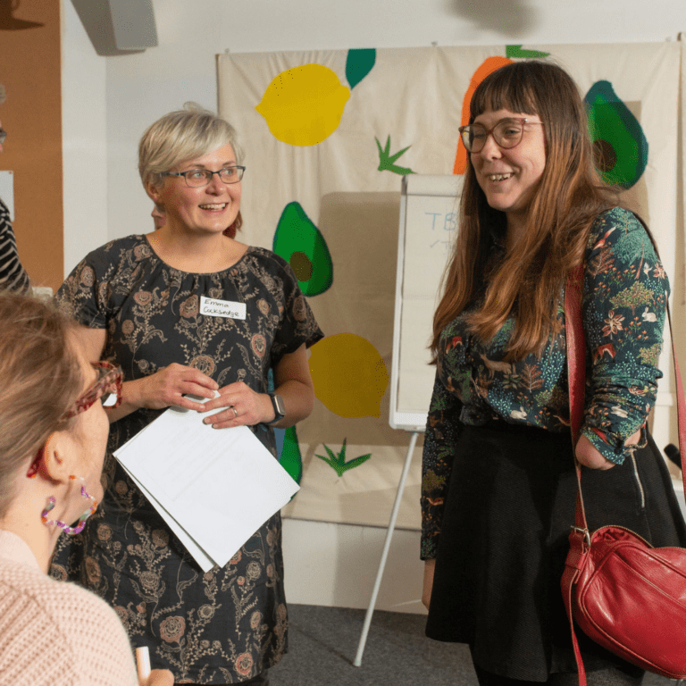 Three people are looking at each other and smiling. Two are standing and facing towards the camera. One is sitting down and facing away from the camera.