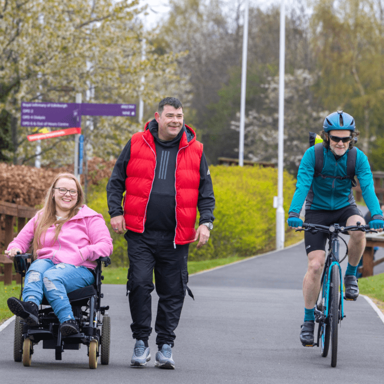 Image shows 3 people on a path. One is a wheelchair user, 1 is walking, 1 is cycling. They look happy. There are trees in the background.