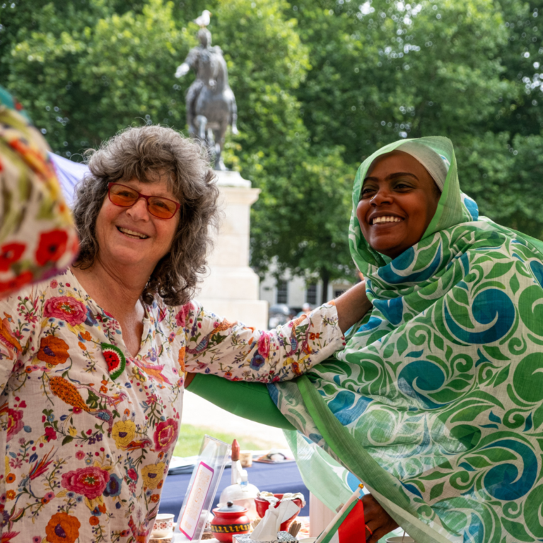 Two people with their arms around each other. They are looking at the camera and smiling. In the background there are some trees.