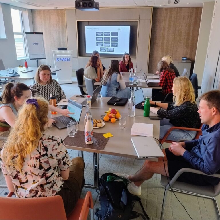 Group of people sitting around a table with laptops in front of a screen