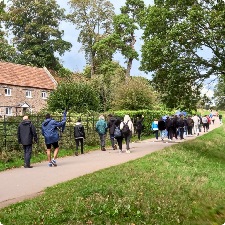 A group of people walking along a path next to a cottage and some trees and grass