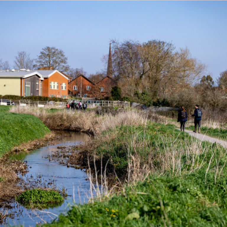 A stream with grassy banks running alongside a winding path leading to a row of houses. There are 2 people walking along the path. The weather is bright.