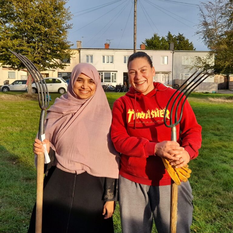 A photo of two people stood next to each other smiling at the camera. They are each holding a pitch fork. They are stood in an urban park in the sunshine.