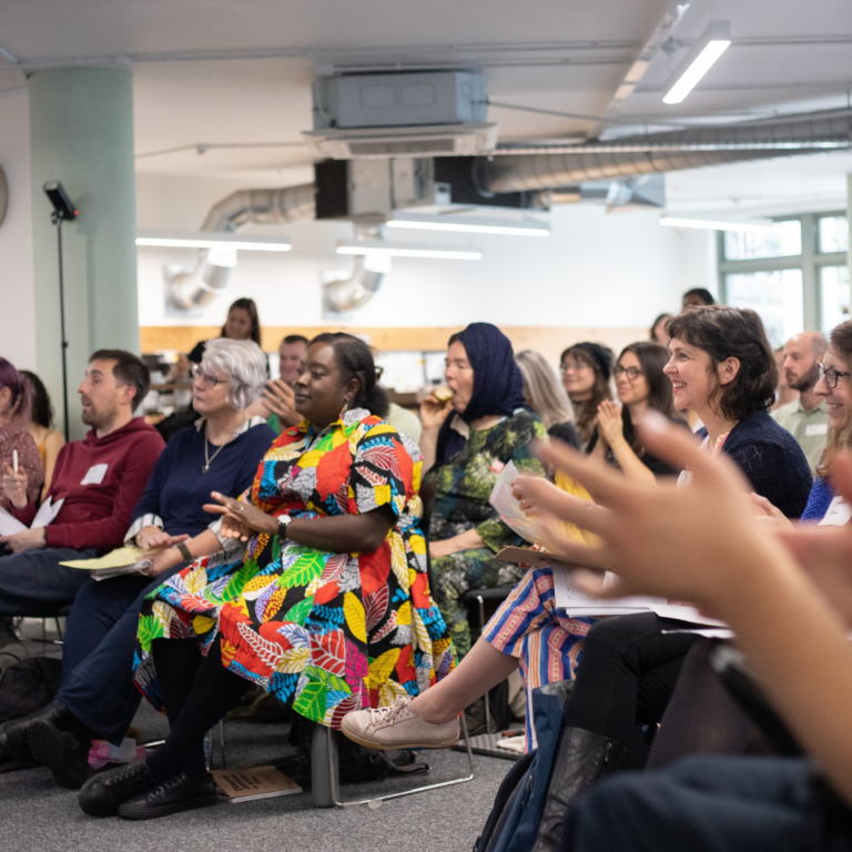 A group of people sat in rows facing the front of a room. They are smiling and clapping.