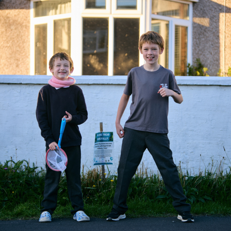 Two young boys smiling. They are stood on grass verge outside a house. There is a sign saying 'please tread carefully'.