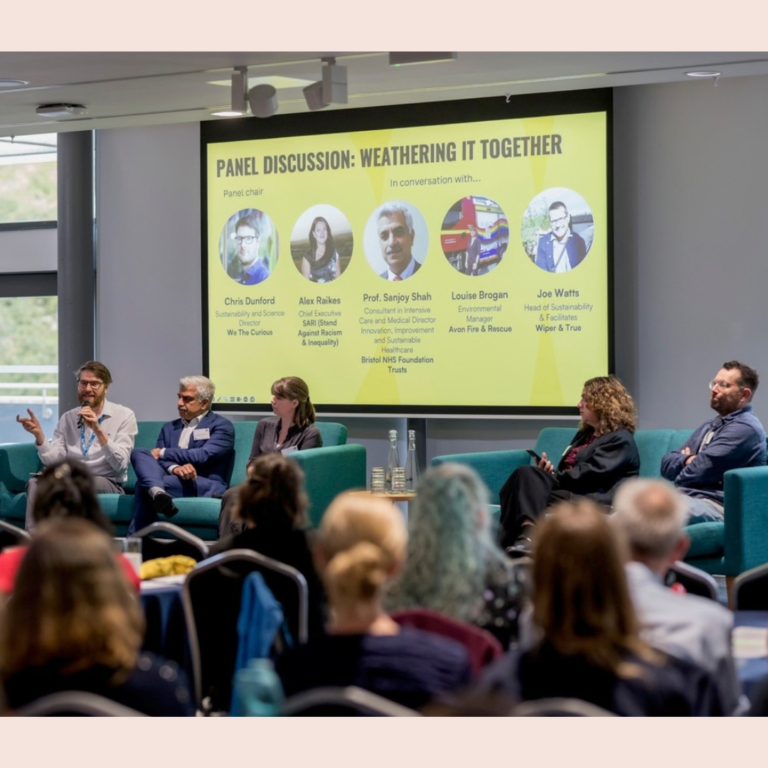 Five people sat on a stage infront of an audience. Behind them is a yellow slide 'PAnel discussion: weathering it together'