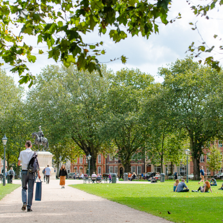 Queens Square with people sat on the grass and walking through the centre. The weather is sunny.